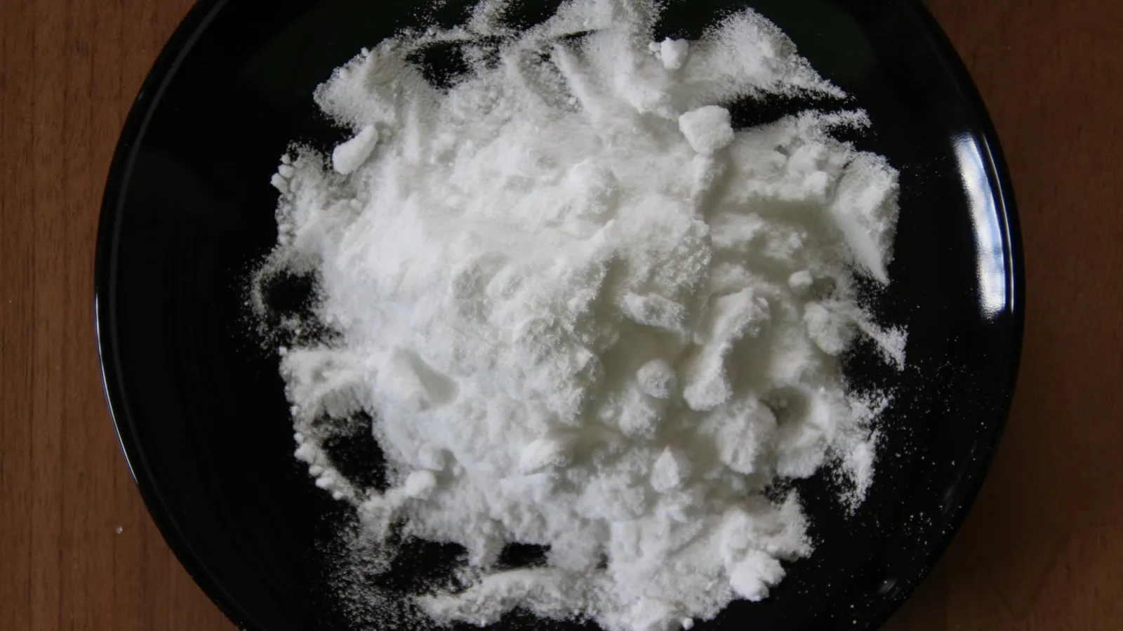 Un tas de carbonate de sodium, poudre blanche en grumeaux, sur une assiette noire posée sur une table en bois
