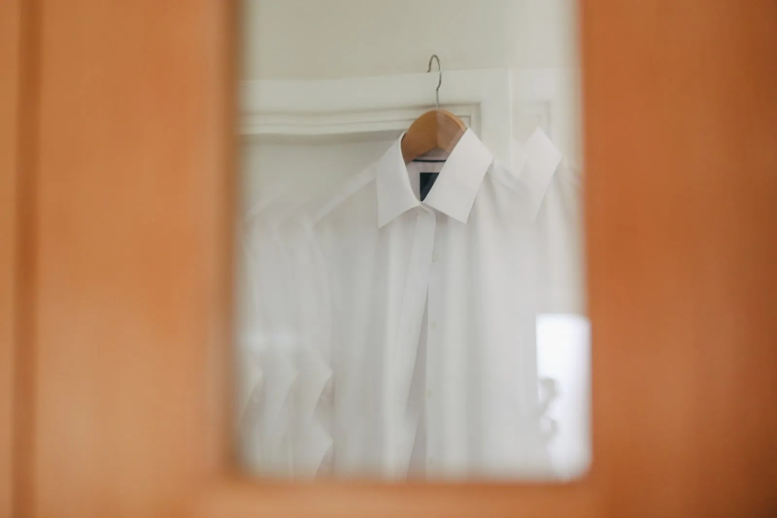 Une chemise blanche à col pointu suspendue à un cintre en bois, vue à travers l’embrasure d’une porte